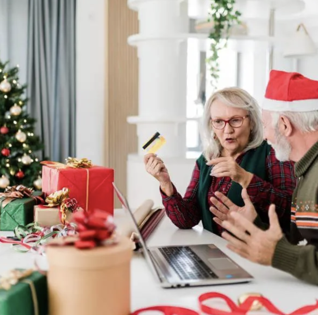 Older man and woman during Christmas season sitting in front of a laptop. The woman is holding a credit card for an online purchase. A decorated Christmas tree is nearby.